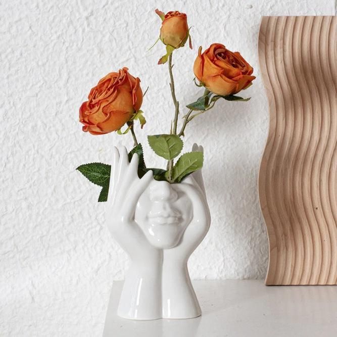 White vase shaped like a face holding orange roses against a white wall.