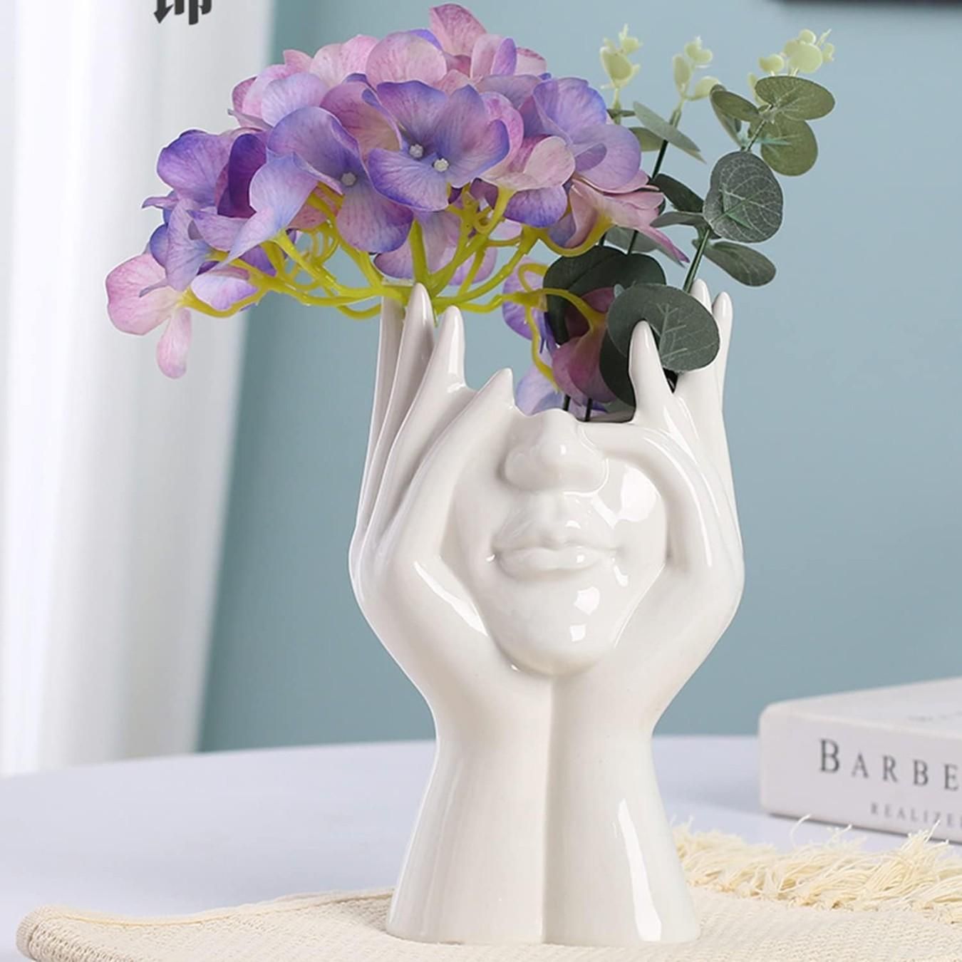 White ceramic vase shaped like a hand holding flowers on a light surface.