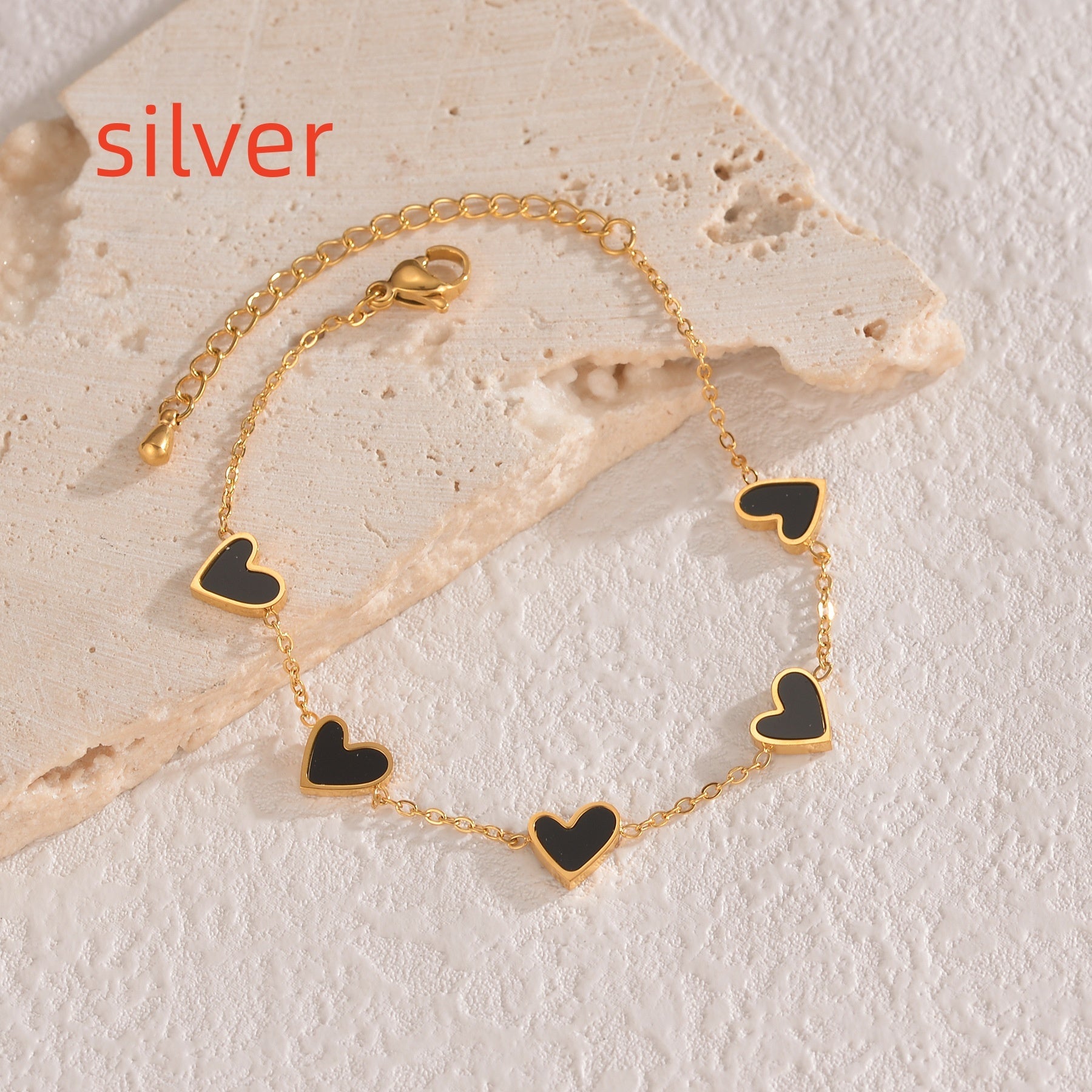 Gold bracelet with black heart charms on a textured stone surface, labeled 'silver'.