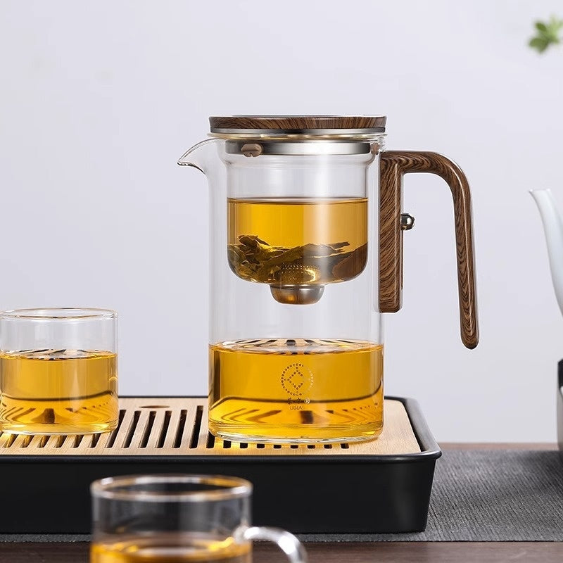 Clear glass teapot with wooden handle and lid, filled with tea, on a wooden tray.