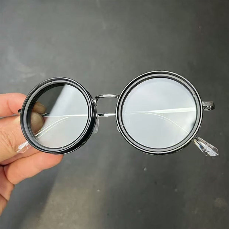 A pair of round sunglasses with a metallic frame held in a hand, viewed from the front with a dark lenses and a visible bridge.