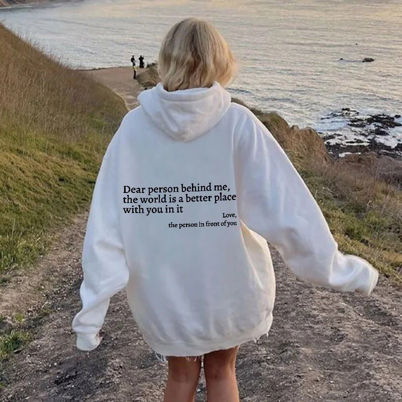 Person wearing a white hoodie with a message on the back, standing on a path by a body of water.