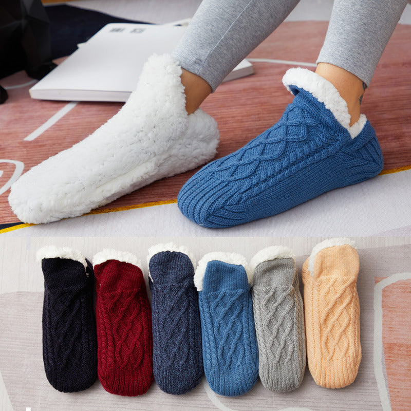 Pair of blue cable knit slippers with fluffy trim worn by a person, with six pairs of similar slippers displayed below on a wooden floor.